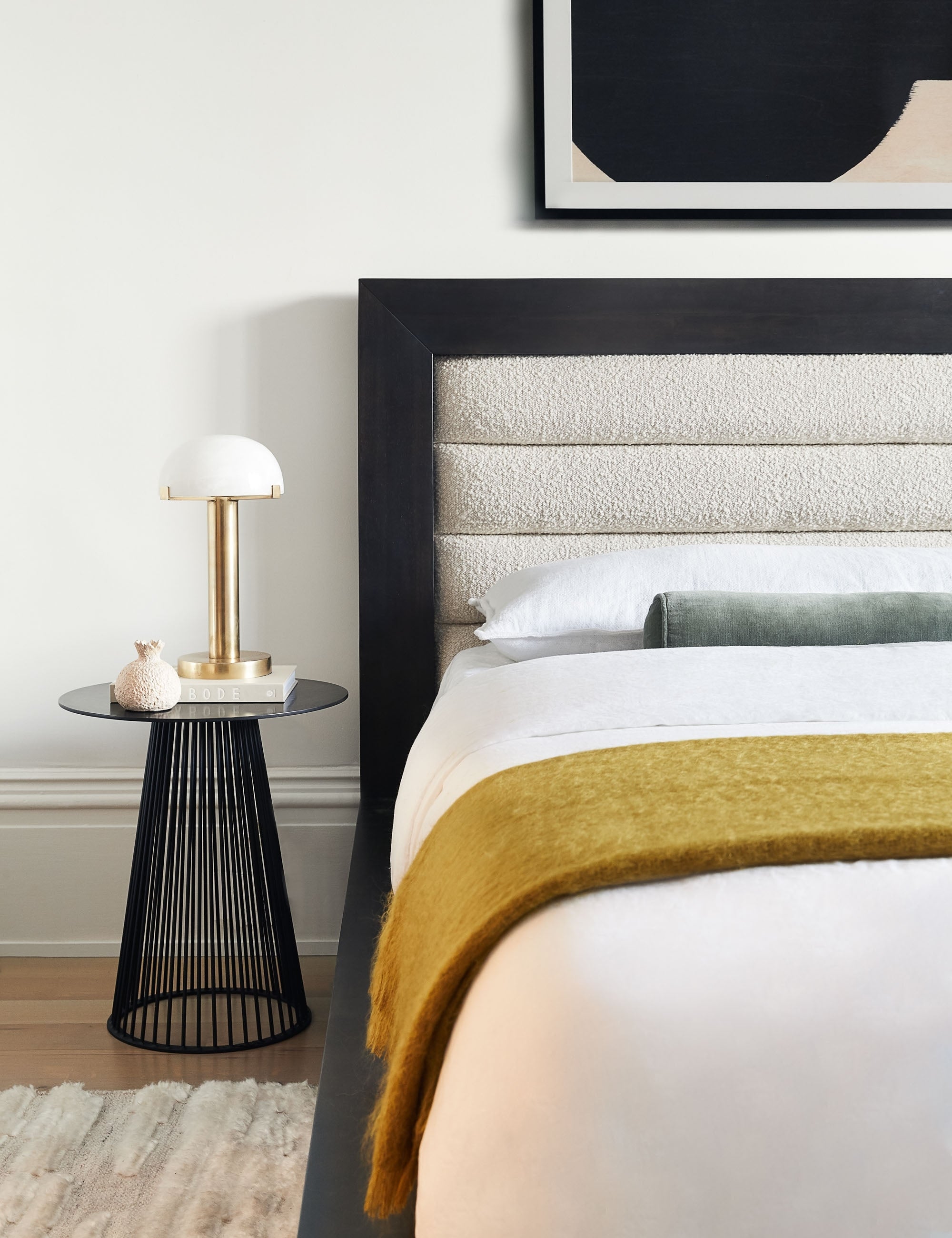 | The Lelani table lamp with half-moon shade on a gold base sits in a bedroom atop a sculptural black nightstand to the left of a bed with a wooden framed boucle headboard