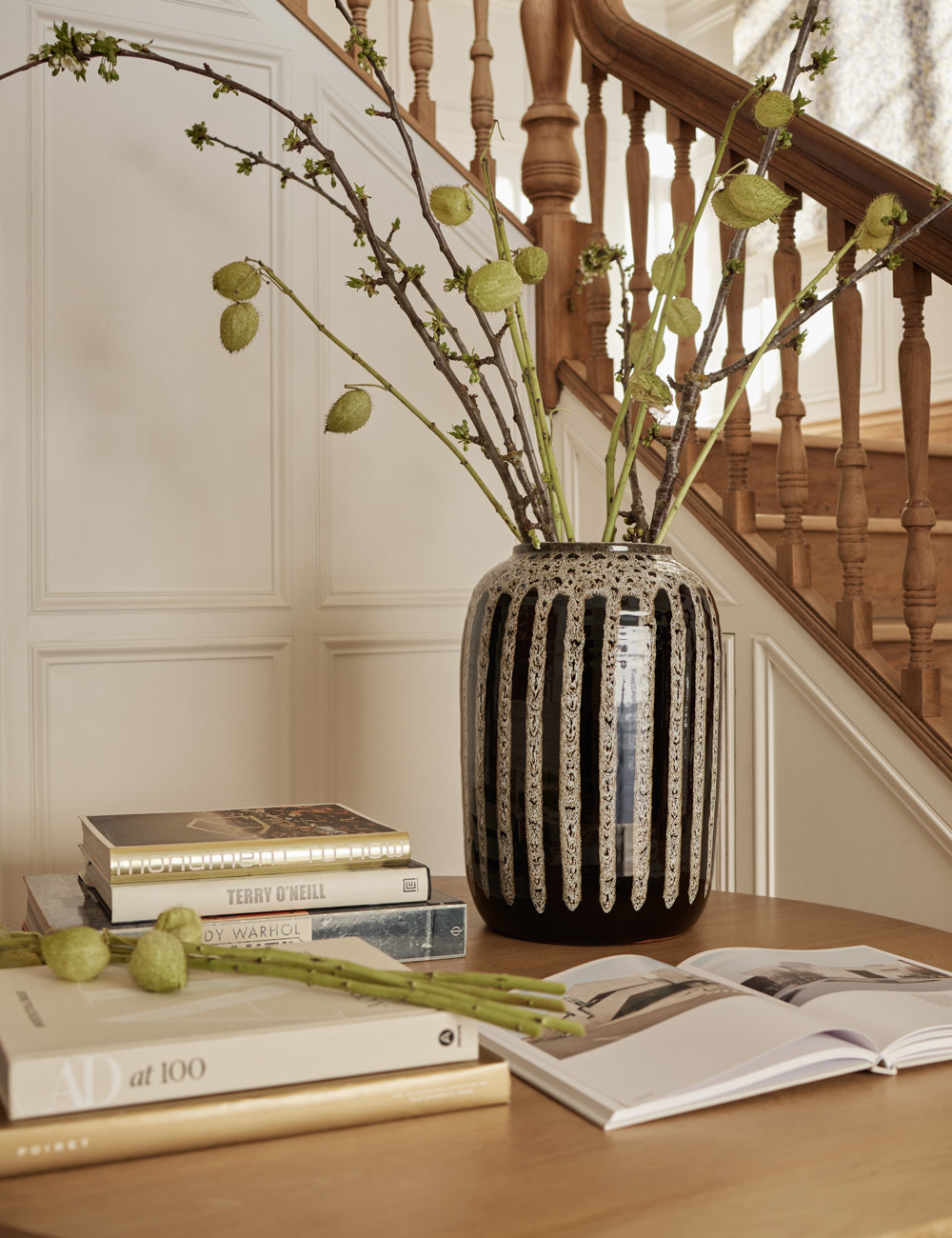 | Wheaton full-bodied black and natural striped vase with a plant inside sits on a table with stacks of books and a staircase in the background